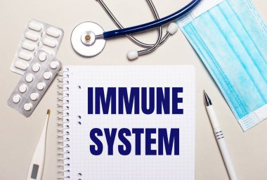 Açık gri bir arka planda, açık mavi tek kullanımlık yüz maskesi, bir stetoskop, bir elektronik termometre, haplar, bir kalem ve üzerinde IMMUNE Sistemleri yazan bir defter. Tıbbi konsept