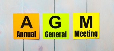 Açık mavi ahşap arka planda AGM Yıllık Genel Toplantısı yazılı üç renkli etiket