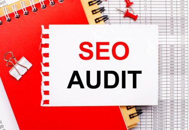 Raporların arka planında kırmızı ve kahverengi not defterleri, beyaz ataç, kırmızı düğmeler ve SEO AUDIT yazılı beyaz bir kağıt var. İş konsepti