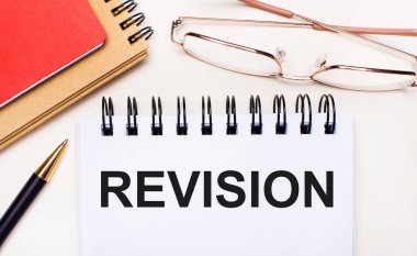 Hafif bir arkaplanda altın çerçeveli gözlük, bir kalem, kahverengi ve kırmızı not defterleri ve üzerinde REVISION yazan beyaz bir defter. İş konsepti