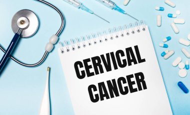 Açık mavi arka planda, bir elektronik termometre, bir steteskop, beyaz ve mavi haplar, şırıngalar ve CERVICAL CANCER yazılı bir defter. Tıbbi konsept