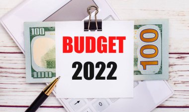 Hafif ahşap bir masanın üzerinde beyaz bir hesap makinesi, bir kalem, faturalar ve siyah bir ataçın altında bir kağıt parçası var.