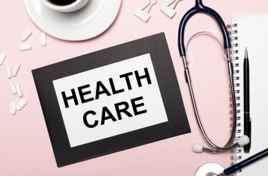 Açık pembe arka planda, kalem, steteskop, beyaz haplar, ataçlar ve HeALTH CARE yazılı bir kağıt olan bir defter. Tıbbi konsept