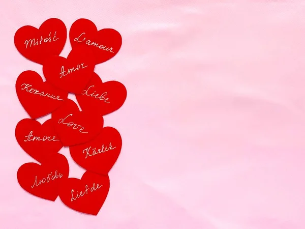 Red paper hearts with white handwritten text. The word love in different languages. Pink fabric, copy space.