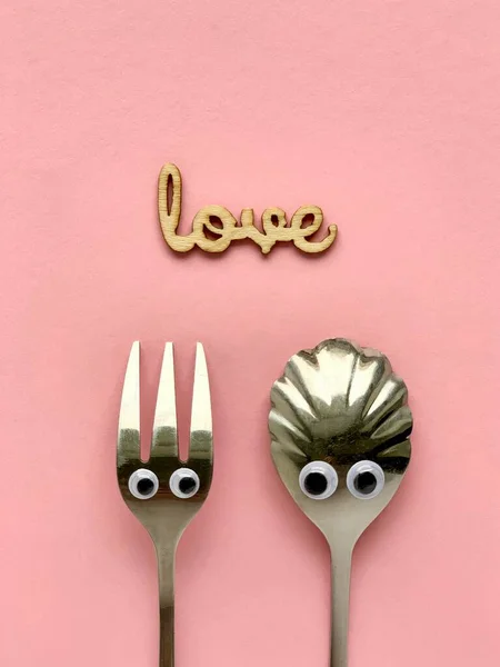 Επιδόρπιο πιρούνι και κουταλάκι του γλυκού με μάτια, ξύλινη λέξη Αγάπη σε ροζ φόντο. Ειδύλλιο. — Φωτογραφία Αρχείου