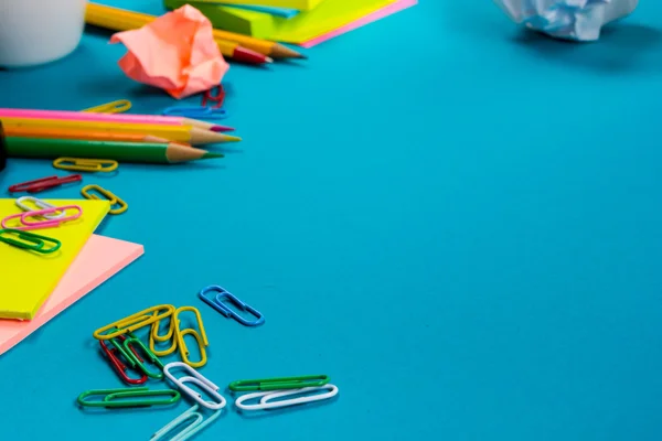 Office table desk with set of colorful supplies, white blank note pad, cup, pen, pc, crumpled paper, flower on blue background. Top view and copy space for text