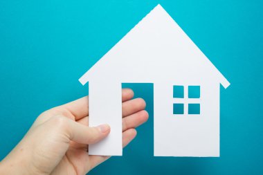 Hand holding white paper house figure on blue background. Real Estate Concept. Ecological building. Copy space top view.