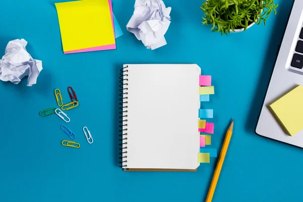 Schreibtisch mit buntem Zubehör, weißem Notizblock, Tasse, Stift, PC, zerknülltem Papier, Blume auf blauem Hintergrund. Ansicht von oben und Kopierraum für Text — Stockfoto