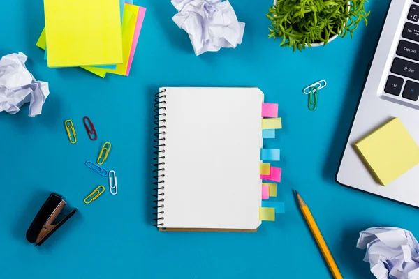 Schreibtisch mit buntem Zubehör, weißem Notizblock, Tasse, Stift, PC, zerknülltem Papier, Blume auf blauem Hintergrund. Ansicht von oben und Kopierraum für Text — Stockfoto