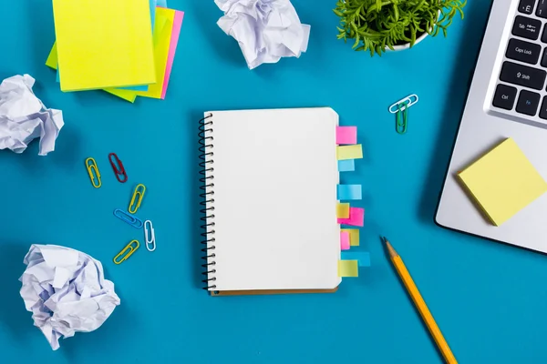 Schreibtisch mit buntem Zubehör, weißem Notizblock, Tasse, Stift, PC, zerknülltem Papier, Blume auf blauem Hintergrund. Ansicht von oben und Kopierraum für Text — Stockfoto