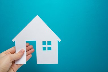 Hand holding white paper house figure on blue background. Real Estate Concept. Ecological building. Copy space top view.