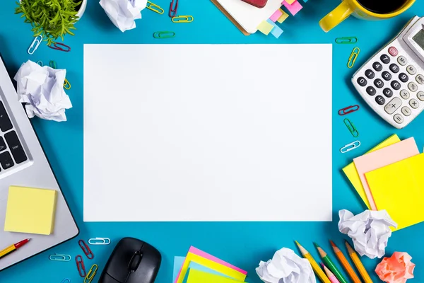 Office table desk with set of colorful supplies, white blank note pad, cup, pen, pc, crumpled paper, flower on blue background. Top view and copy space for text