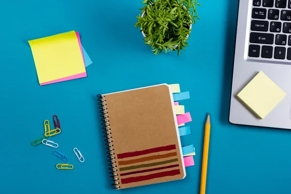 Schreibtisch mit buntem Zubehör, weißem Notizblock, Tasse, Stift, PC, zerknülltem Papier, Blume auf blauem Hintergrund. Ansicht von oben und Kopierraum für Text — Stockfoto