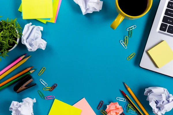 Escritorio de mesa de oficina con conjunto de suministros de colores, bloc de notas blanco, taza, pluma, PC, papel arrugado, flor sobre fondo azul. Vista superior y espacio de copia para texto —  Fotos de Stock