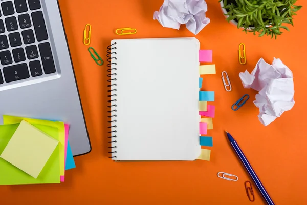 Office table desk with set of colorful supplies, white blank note pad, cup, pen, pc, crumpled paper, flower on orange background. Top view and copy space for text — Φωτογραφία Αρχείου
