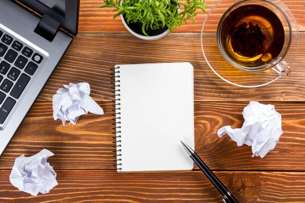 Tabell skrivbord med leveranser, vit tomt anteckningsblock, cup, penna, pc, skrynkligt papper, blomma på trä bakgrund. Ovanifrån — Stockfoto