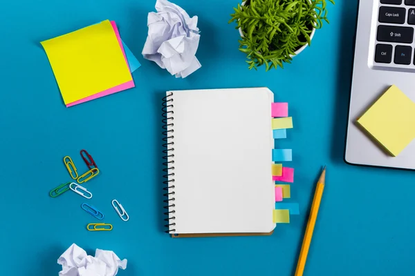 Schreibtisch mit buntem Zubehör, weißem Notizblock, Tasse, Stift, PC, zerknülltem Papier, Blume auf blauem Hintergrund. Ansicht von oben und Kopierraum für Text — Stockfoto