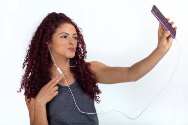 Cep telefonu ve kulaklık kullanan, gülümseyen ve selfie çeken mutlu bir kadının portresi. Beyaz bir arkaplanda izole edilmiş.