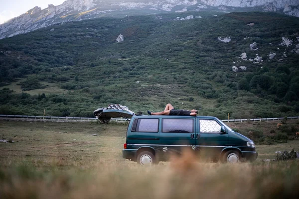 Genç adam doğanın ortasında yeşil karavanının çatısında yatıyor.