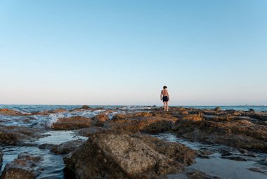 Genç adam gün batımında deniz kenarındaki kayaların üzerinde yürüyor.