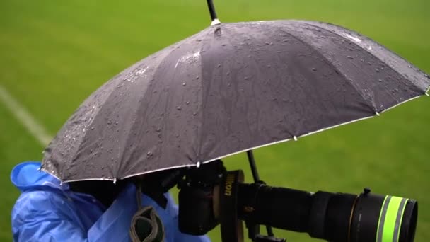 Fotograf in blauem Regenmantel versteckt sich mit schwarzem Regenschirm vor Regen — Stockvideo