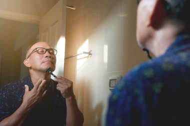 Asyalı yaşlı adam banyoda sakalını tıraş oluyor.