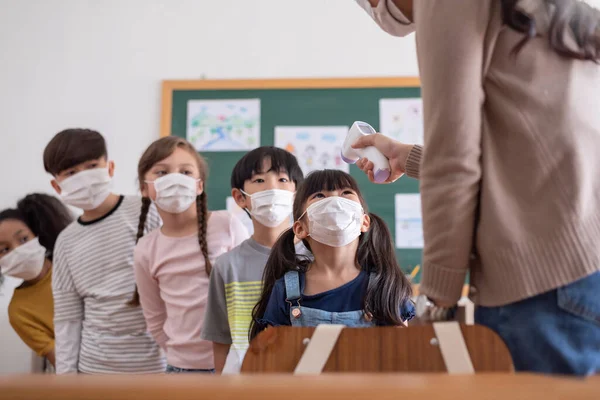 Teacher using digital thermometer for check temperature on Asian girl and kid foreheads. Diversity students wearing protective face mask with COVID-19 Screening in the classroom at Elementary school