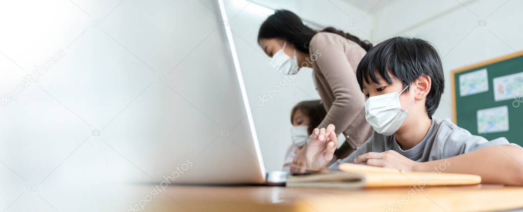 Asian boy wearing protective face mask learning on laptop at desk in classroom. Teacher and Students studying at international school. Education, E-Learning with internet online technology. Banner.