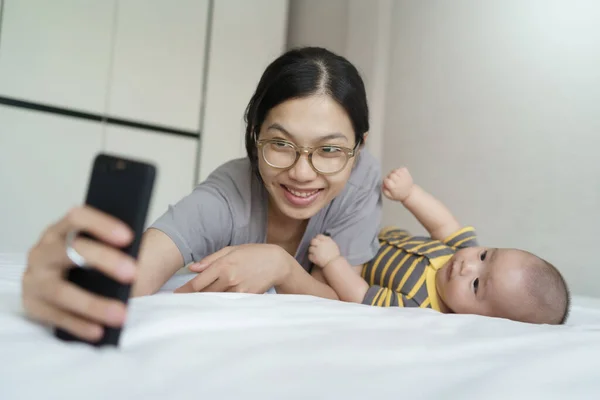 Happy Smiling Asian Young mother and newborn baby boy lying on bed taking selfie with mobile phone together at home. Cheerful Mom and son having fun with smartphone