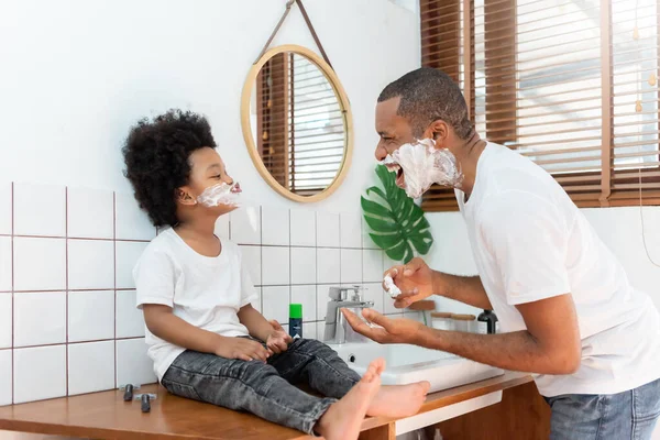 放假的时候 在家里的浴室里 滑稽的黑人爸爸和小男孩一边笑一边刮胡子泡沫 快乐的非洲家庭早上玩得开心 — 图库照片