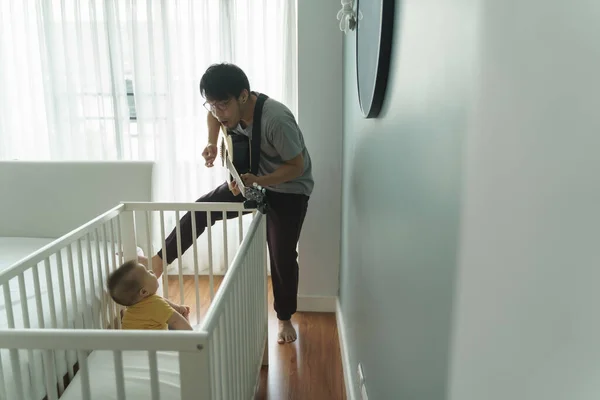 Happy Asian Dad singing and playing acoustic guitar while little adorable baby boy son sitting in the crib. Family having fun and enjoying in the music together. Hobbies and Leisure Lifestyle at home.