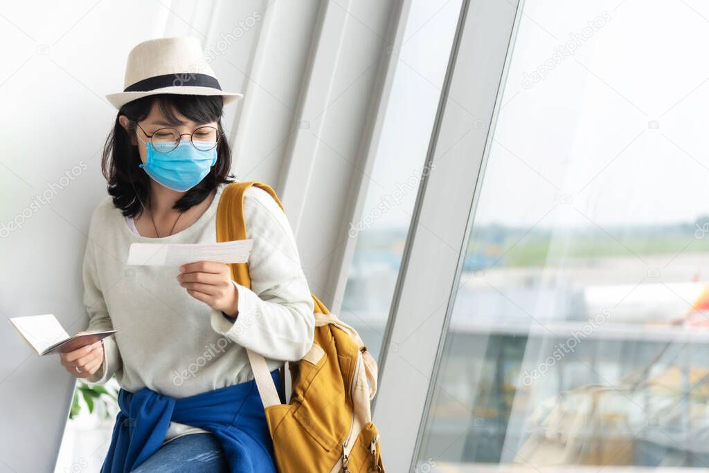 Asian Female tourist wearing protective face mask holding boarding pass waiting for the flight near the window of the airport terminal on her vacation. New normal Lifestyle