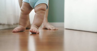 Close up Asian Mother holding her little baby learning to walk on wooden floor at home. Cute toddler enjoying the first steps with mom.