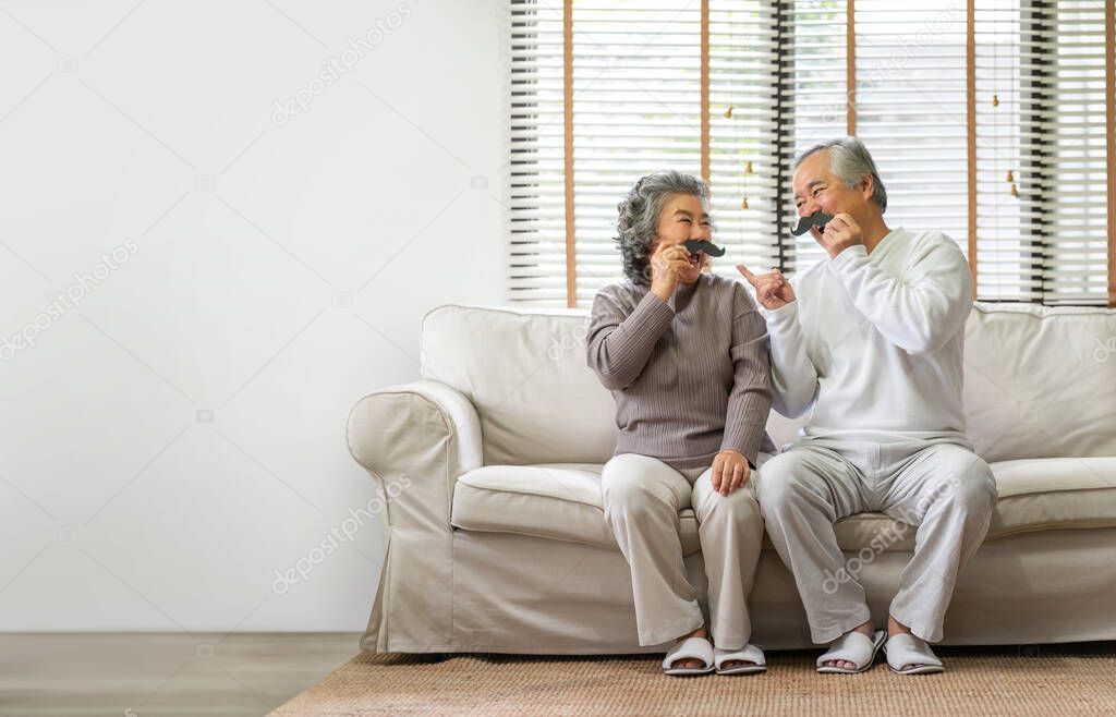 Happy Asian Senior Couple sitting on sofa and playing fake mustaches at home togetherness. 