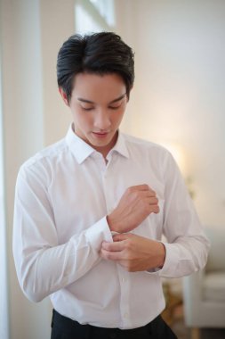 Asyalı damat gömleğini ilikliyor ve düğün törenine hazırlanıyor. Sakin, kendinden emin ve büyük günü için heyecanlı..