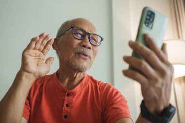 Kırmızı tişörtlü yaşlı Asyalı adam ailesiyle görüntülü konuşma yapmak için akıllı telefon kullanıyor, sevinçle el sallıyor ve evdeki dijital bağlantıyla kendini yakın hissediyor..