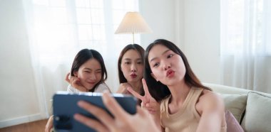 Makyajdan sonra akıllı telefonuyla gülümseyen ve poz veren üç Asyalı genç kadın, evde arkadaşlığın ve mutluluğun tadını çıkarıyorlar..