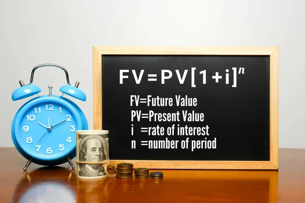 Time value of money formula on blackboard with alarm clock, fake money and coin.