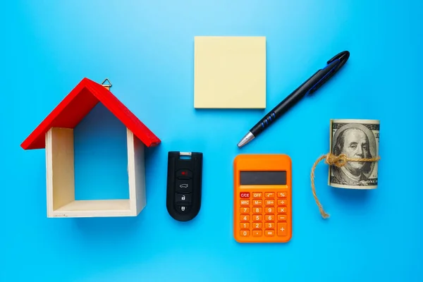A picture house miniature, with fake money, copyspace notepad, car key, calculator, and pen on blue background.