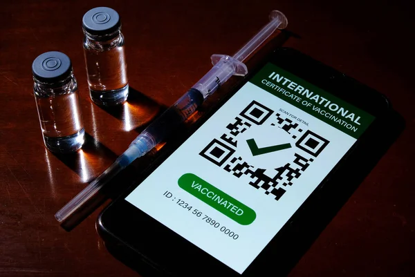 A picture in dark mode of smartphone with fake International Certificate of Vaccination on it, two doses of vaccine and injection syringe insight on the table. Certicate of vaccine will give after complete 2 doses of Covid-19 vaccine