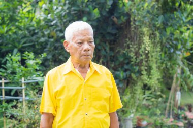 Bahçesinde endişeli bir şekilde duran sarı tişörtlü yaşlı adamın portresi.