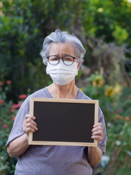 A senior woman standing holding a blank black label and wearing a face mask for protecting covid-19, coronavirus, bacteria, pollen grains, air pollution PM 2.5, and more. Healthcare concept.