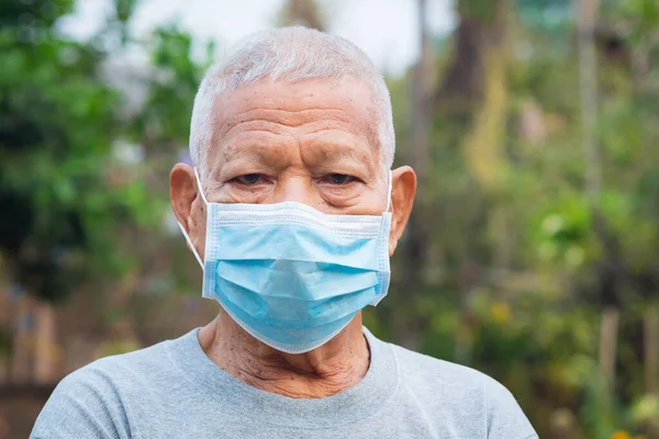 Elderly Asian man wearing a mask and looking at the camera while standing in a garden. Mask for protecting covid-19, coronavirus, and more. Concept of aged people and healthcare.