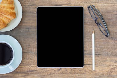 Tablet, kalem, bardaklar, bir fincan kahve ve ahşap bir masaya konmuş kruvasan ile bir masanın üst görüntüsü. Evden çalışıyorum. Metin için boşluk.