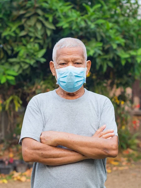 An elderly Asian man wearing a mask and arms crossed while standing in a garden. Mask for protecting covid-19, virus, coronavirus, bacteria, pollen grains. Concept of aged people and healthcare.
