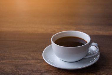 Tahta bir masanın üzerinde güneş ışığıyla birlikte beyaz bir fincan kahve. Mesaj için yer var. Sabah rahatla. Rahatlama kavramı.