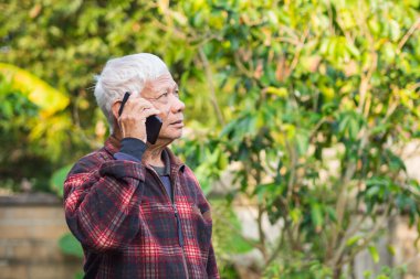 Yaşlı bir adam akıllı telefon kullanıyor ve bahçede dikilirken yukarı bakıyor. Mesaj için yer var. Yaşlılar ve iletişim kavramı.