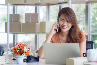 Asyalı iş kadını akıllı telefon kullanıyor ve ofiste otururken dizüstü bilgisayara bakıyor. Genç bayan iş görüşmeleri yapıyor. İş ve e-ticaret kavramı.