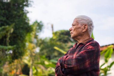 Yaşlı bir adamın kolları çapraz bağlanmış ve bahçede dikilirken yukarı bakan portresi. Mesaj için yer var. Yaşlılar ve sağlık hizmetleri kavramı.