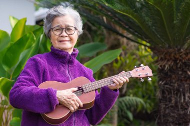 Kısa gri saçlı, gözlüklü ve kapının önünde dikilirken gitar çalan neşeli, yaşlı Asyalı kadın. Yaşlılar ve rahatlama kavramı.